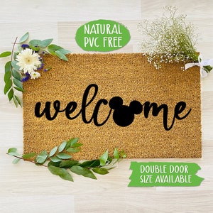 May include: A brown coir doormat with the word "welcome" in black script with a black Mickey Mouse head in the middle of the word. The text "Natural PVC Free" is in a green circle at the top of the image and the text "Double Door Size Available" is in a green rectangle at the bottom of the image.