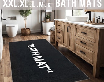 May include: Black bath mat with white text that reads "BATH MAT" in a modern, minimalist style. The mat is placed on a wooden floor in a bathroom setting.