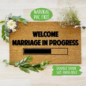 May include: A brown coir doormat with the text "Welcome Marriage in Progress" and a black progress bar underneath. The text "Natural PVC Free" is on a green circle in the top left corner. The text "Double Door Size Available" is on a green rectangle in the bottom right corner.