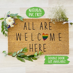 May include: A brown coir doormat with the text "ALL ARE WELCOME HERE" in black lettering. A rainbow heart is in the middle of the word "WELCOME". The text "NATURAL PVC FREE" is in a green circle at the top of the image. The text "DOUBLE DOOR SIZE AVAILABLE" is in a green rectangle at the bottom of the image.