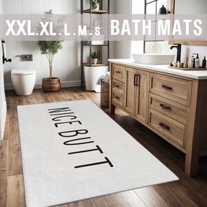 May include: White bath mat with the text "NICE BUTT" printed in black. The mat is in a bathroom with a wooden vanity and a white sink.