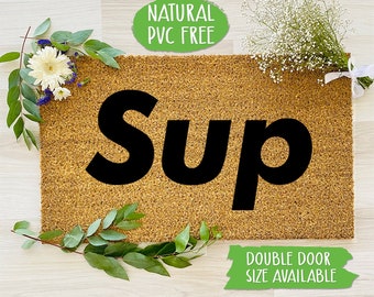 May include: A brown coir doormat with the word "Sup" in black block letters. The doormat is surrounded by greenery and white flowers. A green label reads "Natural PVC Free" and another green label reads "Double Door Size Available".