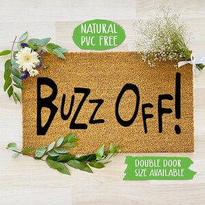 May include: A brown coir doormat with the text "Buzz Off!" in black handwritten font. The doormat is surrounded by greenery and white flowers. The text "Natural PVC Free" is in a green circle above the doormat. The text "Double Door Size Available" is in a green rectangle below the doormat.