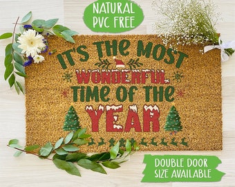 May include: A brown coir doormat with the text "It's the most wonderful time of the year" in red and green lettering. The text is surrounded by a green wreath and snowflakes. The doormat also has a small Christmas tree on each side of the text. The doormat is on a light wood floor. The text "Natural PVC Free" is in a green circle in the top left corner of the image. The text "Double Door Size Available" is in a green rectangle in the bottom right corner of the image.