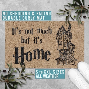 May include: A rectangular doormat with a textured, tan surface. The mat features the phrase "It's not much but it's Home" in black lettering, along with a sketch of a spooky house. The text "NO SHEDDING & FADING DURABLE CURLY MAT" and "S TO XXL SIZES ALL WEATHER" are also present.