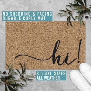 Puede incluir: Una alfombra de coco marrón con la palabra "hi!" escrita en cursiva negra. La alfombra está diseñada para ser duradera y resistente a la intemperie. Está disponible en varios tamaños.