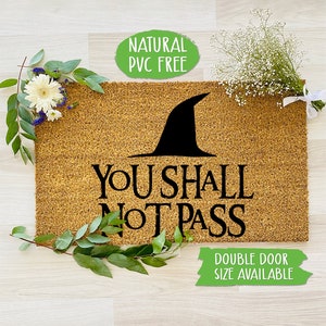 Può includere: Un tappetino per porta in fibra di cocco marrone con il testo "You Shall Not Pass" in lettere nere. Un cappello da strega nero è sopra il testo. Il tappetino è circondato da foglie verdi e fiori bianchi. Un cerchio verde con il testo "Natural PVC Free" è nell'angolo in alto a sinistra. Un rettangolo verde con il testo "Double Door Size Available" è nell'angolo in basso a destra.