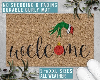May include: A brown coir doormat with the word "welcome" in black cursive lettering. A green Grinch hand with a red Santa hat is holding a red Christmas ornament above the word "welcome". The text "NO SHEDDING & FADING DURABLE CURLY MAT" is at the top of the image. The text "S TO XXL SIZES ALL WEATHER" is at the bottom of the image.