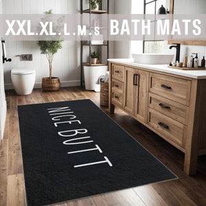 May include: Black bath mat with the text "NICE BUTT" in white lettering. The mat is in a bathroom with a white toilet and a wooden vanity with drawers.