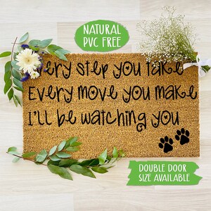 May include: A brown coir doormat with the text "Every step you take Every move you make I'll be watching you" in black cursive font. Two black paw prints are at the bottom of the text. The doormat is on a light wood floor with green plants and white flowers on either side.