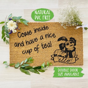 May include: A brown coir doormat with the text "Come inside and have a nice cup of tea!" and a black and white illustration of a creature with the text "ello!" on a banner. The text "NATURAL PVC FREE" is on a green circle above the doormat. The text "DOUBLE DOOR SIZE AVAILABLE" is on a green rectangle below the doormat.
