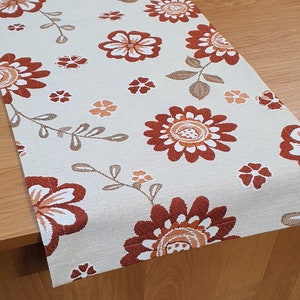 Puede incluir: Camino de mesa color crema con un estampado floral repetido. Las flores son de color óxido, naranja y blanco, con tallos y hojas marrones. El camino de mesa está hecho de tela texturizada y está colocado sobre una mesa de madera.