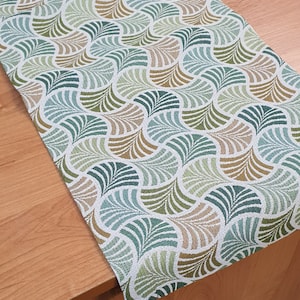 Op de afbeelding: Een groene en bruine tafelrunner met een geometrisch patroon van gestileerde bladeren. De runner is gemaakt van geweven stof en heeft een subtiele glans.