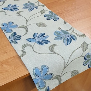 May include: A light blue and white floral table runner with a delicate floral pattern. The runner is made of a soft, textured fabric and features a repeating pattern of blue flowers on a white background.