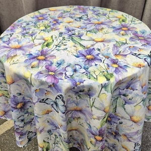 Puede incluir: Mantel redondo con un diseño floral de acuarela con flores moradas y blancas y mariposas. El mantel tiene un fondo claro y es adecuado para una mesa redonda.