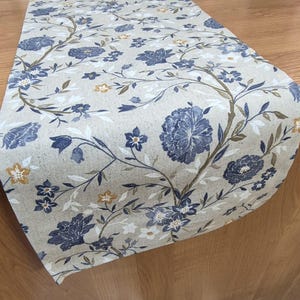 Pode incluir: Um caminho de mesa bege com um padrão floral. O design apresenta flores azuis, brancas e amarelas com caules e galhos marrons. O caminho de mesa é feito de um tecido macio e é drapeado sobre uma superfície de madeira.