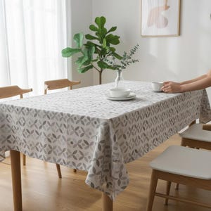 Op de afbeelding: Een rechthoekig tafelkleed met een herhalend grijs bloemmotief bedekt een houten tafel. Witte borden en een kom staan op de tafel. Twee houten stoelen zijn zichtbaar. De handen van een persoon houden een witte beker vast. Een plant en kunstwerk staan op de achtergrond.