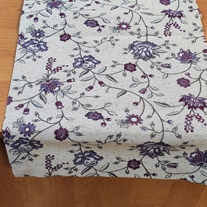 Op de afbeelding: Crèmekleurige tafelloper met een bloemenpatroon. Het ontwerp toont paarse en blauwe bloemen met groene bladeren en stelen. De loper is gemaakt van een textiel met textuur en wordt gepresenteerd op een houten oppervlak.