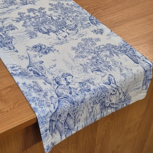 Puede incluir: Un camino de mesa azul y blanco con un patrón toile que representa una escena pastoral con personas, árboles y un edificio. El camino de mesa está sobre una mesa de madera.