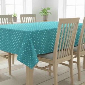 Op de afbeelding: Een turquoise tafelkleed met een wit stippenpatroon bedekt een houten tafel. De tafel wordt omringd door vier houten stoelen. De kamer heeft een neutrale kleurstelling met natuurlijk licht dat van de ramen komt.