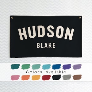 May include: Black canvas banner with white text that reads "HUDSON BLAKE".  The banner is shown hanging on a white wall.  A color chart below the banner shows 12 different color options for the banner.