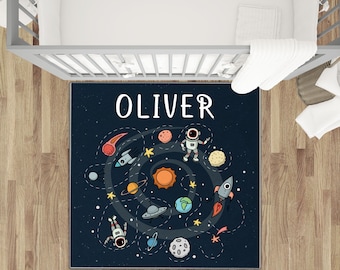 May include: A navy blue rug with a space-themed design, featuring planets, rockets, and astronauts. The name "OLIVER" is displayed in white block letters. The rug is placed in a nursery setting.