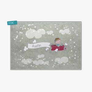 May include: A light gray rug with a white background featuring a cartoon illustration of a boy flying a red airplane with a banner that says "Austin" in white letters. The background is filled with white stars and clouds.