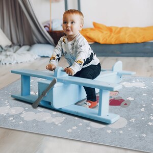 May include: A child sits on a blue toy airplane on a blue and white play rug with a cloud and star pattern.