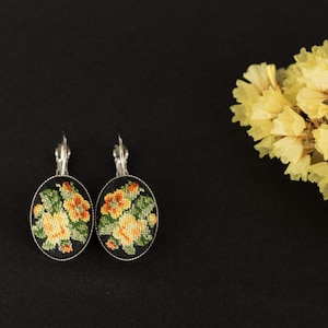 Könnte beinhalten: Ein Paar ovale Ohrringe mit Blumenmuster. Die Ohrringe sind silberfarben mit schwarzem Hintergrund und zeigen gestickte gelbe und grüne Blumen.