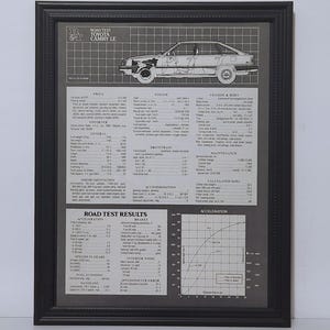 May include: A framed black and white technical print titled Road Test Toyota Camry LE, featuring a side-profile line drawing of the car, detailed engine and chassis specifications, performance data, and an acceleration graph on a gray background.