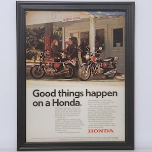May include: Framed vintage Honda motorcycle advertisement. The image features two motorcycles parked in front of a gas station. The advertisement includes the text "Good things happen on a Honda."