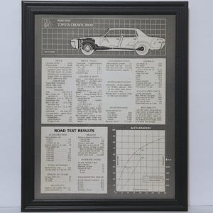 May include: A framed black and white technical print featuring a Toyota Crown 2600 road test report. The document includes a line drawing of the car, detailed specifications, performance data, and an acceleration graph on a grey background.