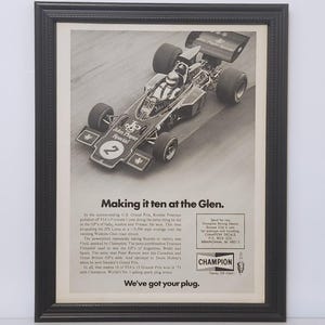 May include: Framed black and white advertisement featuring a Formula 1 race car with the number 2. The ad includes text about the race and the Champion spark plug brand. The text reads "Making it ten at the Glen."