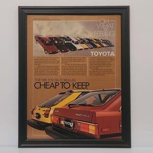 May include: Framed vintage advertisement for the 1981 Toyota Corolla. The ad features several Corolla models in various colors, with the text "Cheap to Keep" and "Oh What a Feeling" prominently displayed. The frame is black.