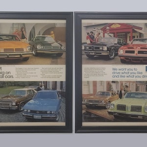 May include: Two framed vintage GM car advertisements. One features the text "GM is big on small cars." The other reads "We want you to drive what you like and like what you drive." The ads showcase various classic car models.