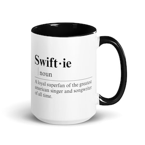 May include: White coffee mug with a black interior and handle. The mug has the text "Swift.ie" with the definition "A loyal superfan of the greatest american singer and songwriter of all time."