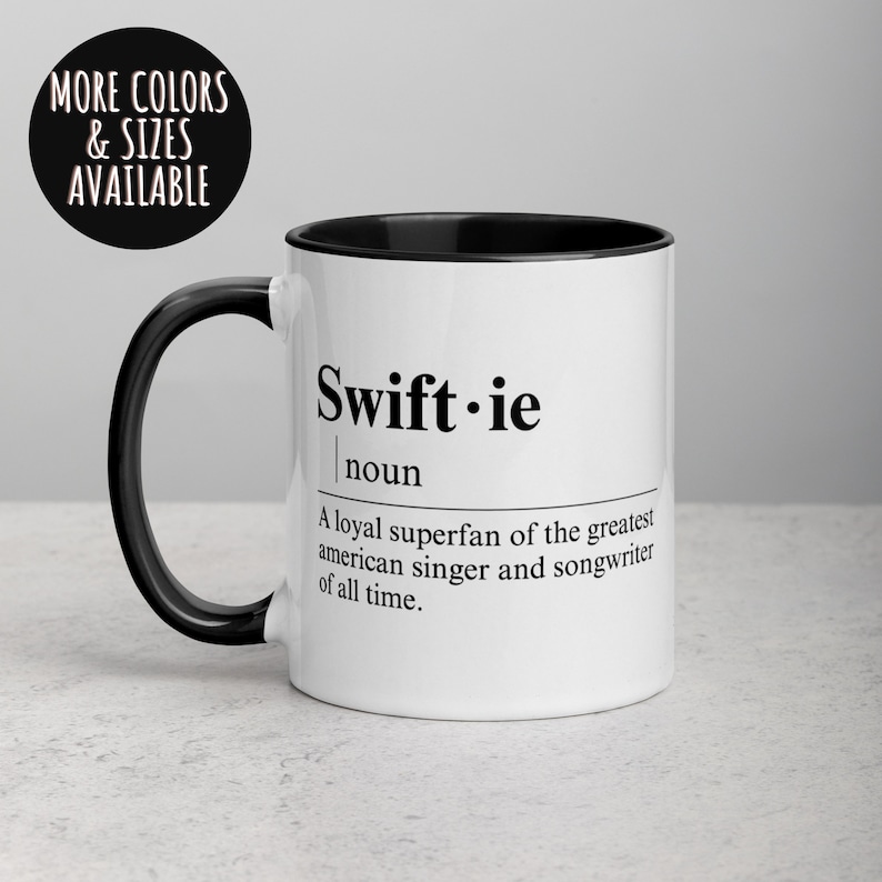 May include: White ceramic coffee mug with a black rim and handle. The mug features black text that reads "Swift-ie" and defines the word as "A loyal superfan of the greatest american singer and songwriter of all time."