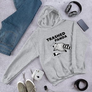 May include: A light grey hoodie featuring the words "Trashed Panda" and a graphic of a raccoon lying near a bottle. The hoodie has a hood and long sleeves. The image also includes blue jeans, headphones, and a smartphone.