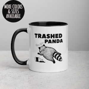 May include: White ceramic mug with a black handle and rim. The mug features the text "Trashed Panda" above a black and white illustration of a raccoon and a spilled bottle. A circular graphic at the top left says "More Colors & Sizes Available."