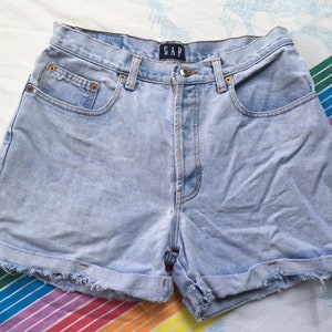May include: Light blue denim shorts with a frayed hem and a cuffed leg. The shorts have a button and zipper closure and a small pocket on the front. The shorts have the word "GAP" written on the back waistband.