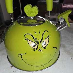 Puede incluir: Una tetera de color verde lima con un gráfico de la cara del Grinch en la parte frontal. El asa de la tapa tiene forma de corazón con el texto The Grinch en relieve. La tetera tiene un asa a juego en verde lima y un pico de metal plateado.