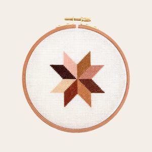 May include: A white embroidery hoop with a brown wooden frame. The hoop contains a cross-stitch design of a starburst with eight points in shades of brown, pink, and beige.