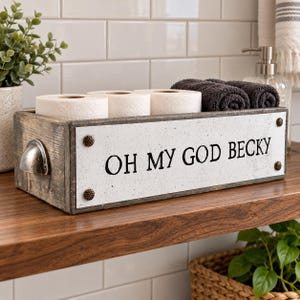 May include: A rectangular wooden storage box with a distressed finish, featuring a white front panel with the black text OH MY GOD BECKY. The box has metal side handles and four decorative studs, holding two rolls of toilet paper and two towels.