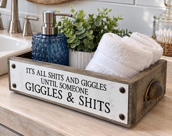 Może przedstawiać: Prostokątna drewniana taca łazienkowa z białą metalową płytką przednią z czarnym napisem: IT'S ALL SHITS AND GIGGLES UNTIL SOMEONE GIGGLES & SHITS. Taca posiada dwa boczne uchwyty wykonane z metalu w kolorze brązu oraz cztery ozdobne nity.