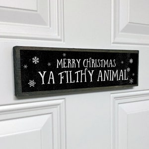May include: A black wooden sign with white text that reads "Merry Christmas Ya Filthy Animal". The sign is decorated with white snowflakes.