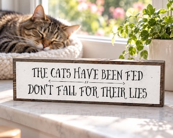 May include: A rectangular wooden sign with a white face and dark brown edges. The text in black reads: THE CATS HAVE BEEN FED, followed by a horizontal line with arrows, and the text: DON'T FALL FOR THEIR LIES.