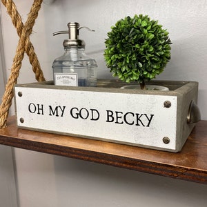 May include: A white wooden box with the text "OH MY GOD BECKY" on the front. The box is sitting on a wooden shelf and contains a clear glass bottle with a pump dispenser and a small potted green plant.