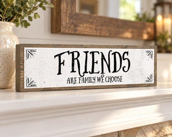 May include: A rectangular wooden sign with a dark brown frame and a white center panel. The text reads FRIENDS ARE FAMILY WE CHOOSE in black lettering. Decorative black corner accents are present on the left and right sides of the sign.