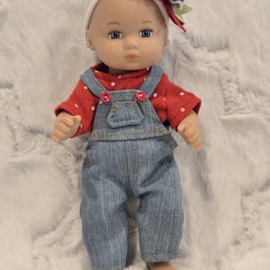 May include: A doll wearing a red polka dot long-sleeve shirt and denim overalls. The doll has blue eyes, a white headband, and a decorative bow. The overalls have red buttons.