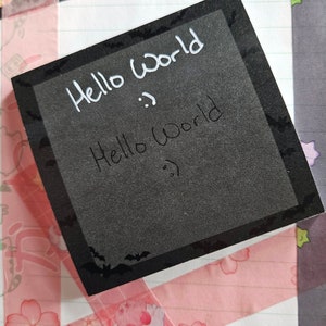 Puede incluir: Una nota adhesiva negra con un borde blanco y siluetas de murciélagos. La nota dice "Hello World" dos veces en escritura blanca.